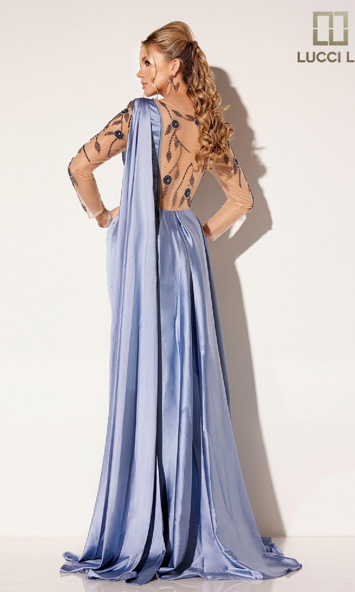 A woman faces away from the camera in the Lucci Lu C8066 long formal dress, featuring periwinkle blue fabric, sheer embroidered back and sleeves, and floral beading, set against a plain background.